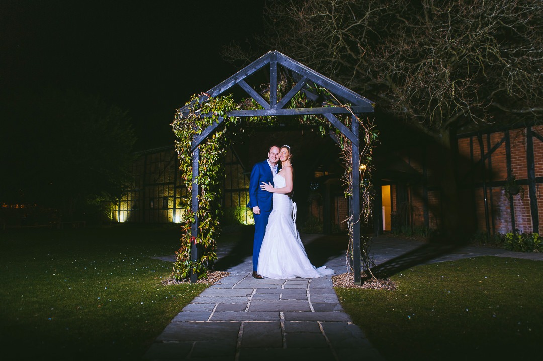 Portrait of Bride and Groom stanidng under a pergola at night-time infront of the Barns Hotel Bedford.